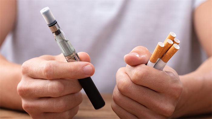 Image of a person in a white t-shirt holding four cigarettes in one hand and a vape device in the other hand. Choose vaping over smoking and it will save your life.